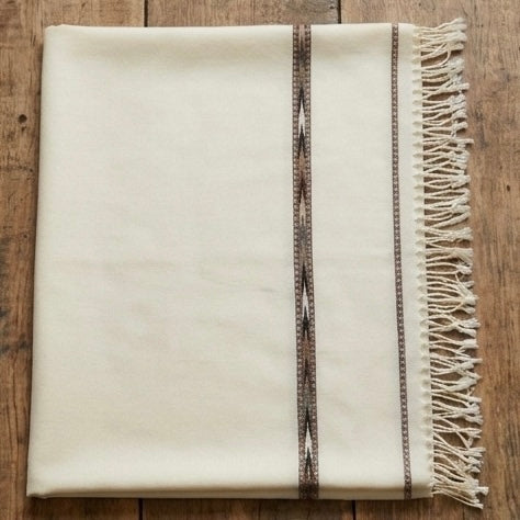 An off-white woven throw blanket with fringe on one side and two vertical decorative borders, laid flat on a rustic wooden surface.
